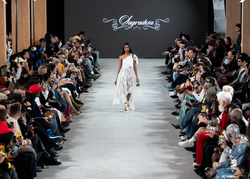 model in white dress walking down a runway with people sitting on the sides